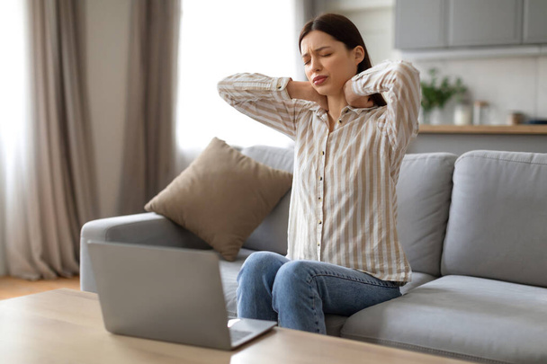 Уставшая молодая женщина массирует шею, демонстрирует признаки напряжения и дискомфорта во время работы из дома, сидя перед ноутбуком на диване - Фото, изображение