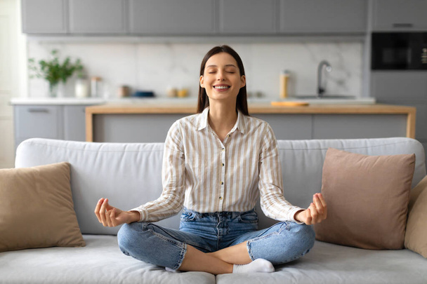 Klidná mladá žena v neformální pruhované košili sedí se zkříženýma nohama, cvičí meditaci na pohodlné šedé pohovce v dobře osvětleném interiéru obývacího pokoje, volný prostor - Fotografie, Obrázek