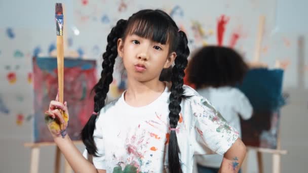 Creative cute girl pose at camera while diverse children painting behind. Young beautiful child looking at camera while standing at stained room and holding paintbrush. Creative activity. Erudition. - Footage, Video