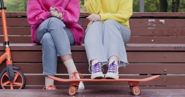 Chica se sienta en tribuna con los pies en el monopatín cerca de amigo con scooter. Mejores amigas se reúnen en el parque de skate para pasar tiempo libre juntas - Metraje, vídeo