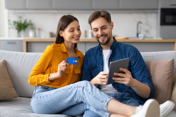 Neşeli genç çift tabletle takılıyor, kredi kartı tutan kadın online alışveriş veya rezervasyon yaptıklarını öne sürüyor, kanepede oturuyorlar. - Fotoğraf, Görsel