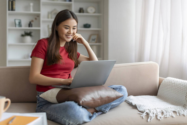 Glückliche junge Asiatin, die zu Hause Filme online auf dem Laptop anschaut, fröhliche Koreanerin, die es sich auf der bequemen Couch mit dem Computer bequem macht, moderne Technologien für ihre Freizeit nutzt und häusliche Pastime genießt - Foto, Bild