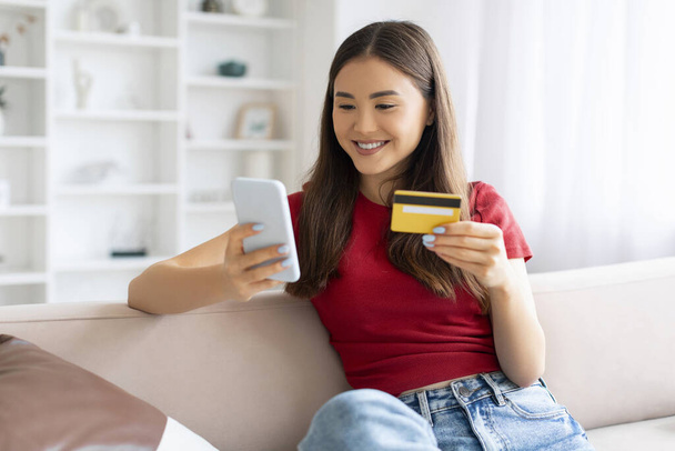 Mobile Payments. Young Beautiful Asian Woman Using Smartphone And Credit Card At Home, Smiling Korean Female Sitting On Couch And Browsing App For E-Commerce, Making Online Shopping, Copy Space - Photo, Image