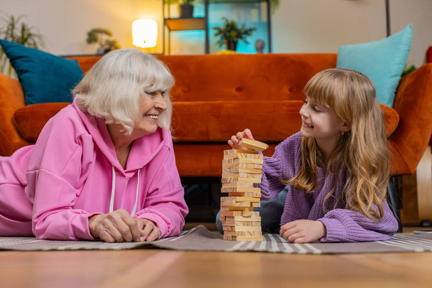 Nadšený malý roztomilý dívka vnučka hrát stavební blok hru s úsměvem starší babička prarodiče. Budování dřevěné věže a odstraňování bloků v obývacím pokoji doma o víkendech. - Fotografie, Obrázek