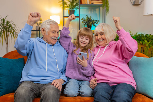 Έκπληξη Καυκάσιος παππούς, γιαγιά και εγγονή χρησιμοποιώντας smartphone, ενώ γιορτάζει την επιτυχία στο σπίτι. Ευτυχισμένο κορίτσι με παππούδες έκπληκτος από online παιχνίδι κερδίσει κάθεται στον καναπέ στο σαλόνι. - Φωτογραφία, εικόνα