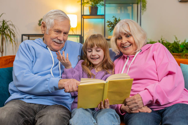 Кавказские бабушка с дедушкой и внучка читают интересную книгу вместе, сидя на диване в гостиной дома. Девушка с бабушкой и дедушкой проводит досуг на удобном диване в квартире - Фото, изображение