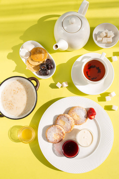 Savor a delightful breakfast with cottage cheese pancakes, creamy oatmeal, aromatic tea, golden honey, jam, and fresh fruit on a sunny yellow table, creating a vibrant start to your day. - Photo, Image
