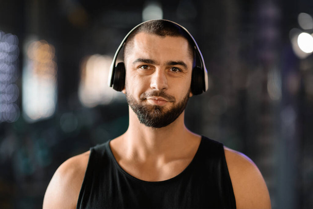 Portrait Of Smiling Athletic Young Man Wearing Wireless Headphones Posing In Gym, Handsome Caucasian Male Listening Music While Training In Sport Club, Enjoying Playlist For Workout, Copy Space - Photo, Image