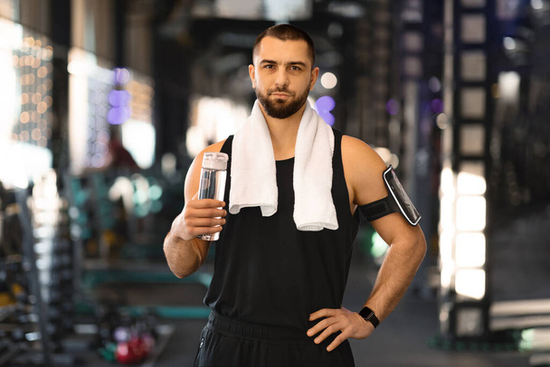 Portret przystojnego młodego sportowca z butelką wody w rękach pozowanie na siłowni podczas odpoczynku po treningu, mięśniowy facet Millennial z ręcznikiem na szyi relaks po treningu, patrząc przed kamerą - Zdjęcie, obraz