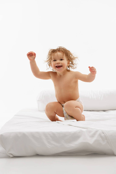 Portrait of charming baby, little girl with curly hair with laugh, jumping on bed in light, airy bedroom against white studio background. Concept of beauty, childhood, motherhood, life, birth. Ad - Photo, Image