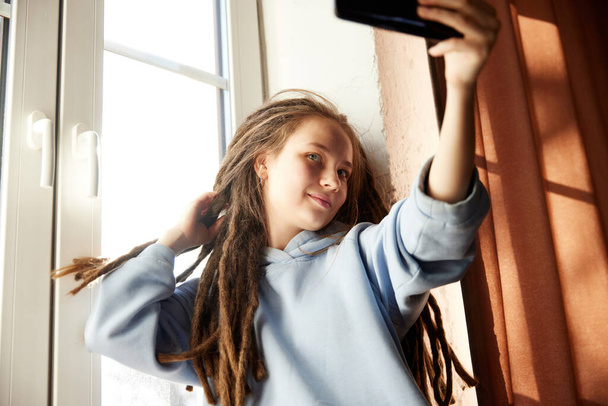 Młoda uśmiechnięta dama z długimi dredami siedzi przy oknie i robi sobie selfie przez telefon, ciesząc się spokojną chwilą. Pojęcie hobby, stylu życia, wolnego czasu, wyrażania siebie, mody i stylu. Ogłoszenie - Zdjęcie, obraz