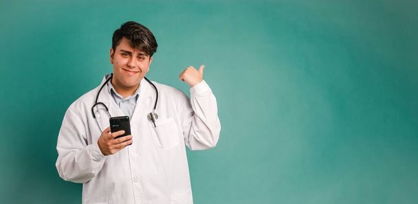 Retrato de un joven médico varón sosteniendo un teléfono inteligente mientras señala el espacio de maqueta sobre un fondo verde - Foto, imagen