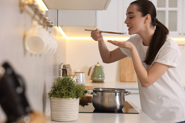 Smiling woman with wooden spoon tasting soup in kitchen - Photo, Image