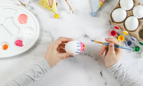 Kafkasyalı küçük bir kız paskalya yumurtasını akrilik boya mavi çizgili bir fırçayla boyuyor, mermer bir masada oturuyor ve paskalya bayramı için bir palet hazırlıyor, düz yakın plan. Şey... - Fotoğraf, Görsel