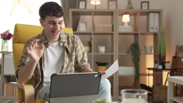 молодий чоловік, білий підліток сидить вдома, використовує цифровий планшет, показує документи листів або результати стипендійних іспитів мають відеодзвінок вдома щаслива посмішка - Кадри, відео