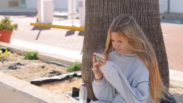 10代の少女が公園に座り,彼女の手に電話をかけている. ティーンエイジャーが携帯電話の画面を横切って指を動かしている. 世代アルファ,世代Z,多様性の概念。 高品質のフルHD映像 - 映像、動画