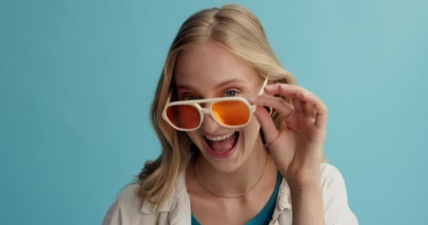 Woman, face and crazy with sunglasses in studio, happy and playful with protection from sun and eye care. Funky, goofy model in portrait and fashion frame with fun and retro style on blue background. - Footage, Video
