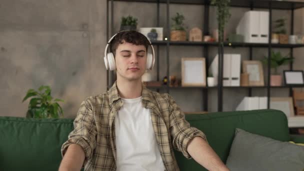 Ένας έφηβος άνδρας νεαρός καυκάσιος άντρας χρησιμοποιεί ακουστικά για online καθοδηγούμενη πρακτική διαλογισμού mindfulness yoga με τα μάτια κλειστά στο σπίτι πραγματικοί άνθρωποι αυτο-φροντίδα εκδήλωση έννοια αντίγραφο χώρο γενιά Z - Πλάνα, βίντεο