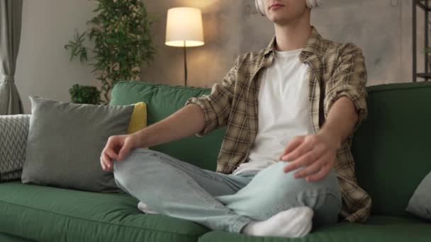 Ένας έφηβος άνδρας νεαρός καυκάσιος άντρας χρησιμοποιεί ακουστικά για online καθοδηγούμενη πρακτική διαλογισμού mindfulness yoga με τα μάτια κλειστά στο σπίτι πραγματικοί άνθρωποι αυτο-φροντίδα εκδήλωση έννοια αντίγραφο χώρο γενιά Z - Πλάνα, βίντεο