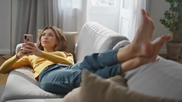 Álmodozó lány pihentető mobiltelefon kényelmes kanapén. Komoly hűsítő nő veszi fel az üzenetrögzítőt a nappaliban. Nyugodt hölgy néz félre fekve puha kanapén. Technológiák és emberek - Felvétel, videó