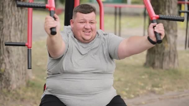 Een dikke jongeman in een grijs t-shirt doet buiten oefeningen op een fitnessapparaat. Worstelen met overgewicht. - Video