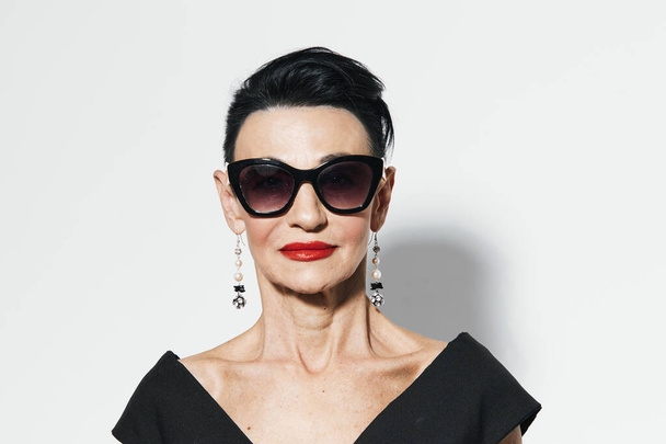 Elegant woman with black hair and red lipstick standing in front of white wall, wearing black dress and red earrings - Photo, Image