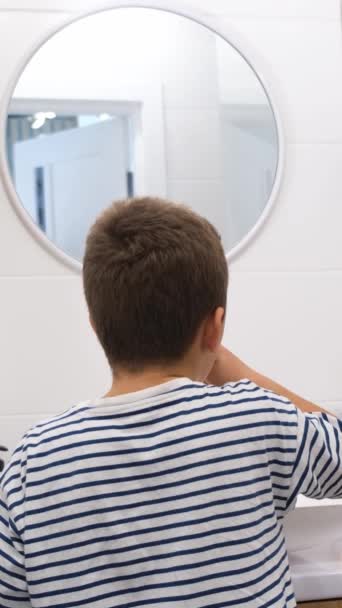 Egy fiatal fiú egy ujját használja, hogy fogat mosson egy kör alakú tükör előtt, egy fémfoglalattal és egy mintás üveggel teli szobában.. - Felvétel, videó