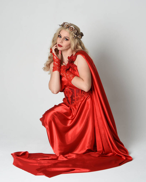 Portrait de pleine longueur d'un beau modèle blond habillé en ancienne déesse mythologique fantaisie en robe de toge de soie rouge fluide, couronne. Pose à genoux assise sur le sol. isolé sur fond de studio blanc. - Photo, image