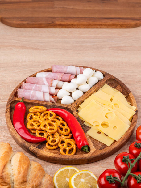 Деревянное блюдо с разнообразными сырами, сосисками и овощами. Сырная тарелка, закуски для праздничного стола. - Фото, изображение
