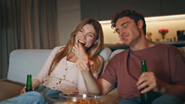 Śmiejąca się para oglądająca sitcom siedzący w przytulnym mieszkaniu w nocy. Szczęśliwi kochankowie jedzący przekąski pijący piwo wieczorem. Uśmiechnięci weseli małżonkowie korzystający z programu telewizyjnego relaks razem na kanapie - Materiał filmowy, wideo