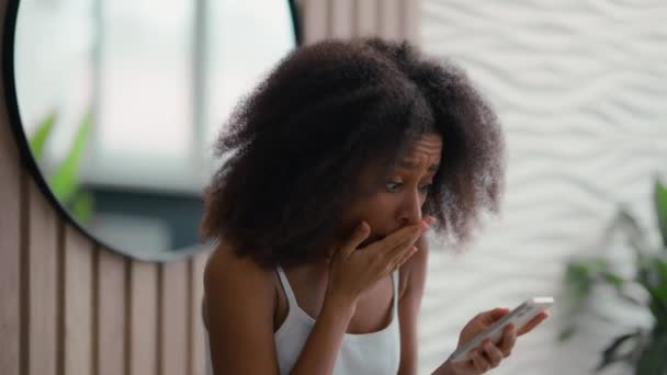 African American displeased young woman in home bathroom bath looking at smartphone screen dissatisfied with bad news message spam scam sms ethnic biracial girl unhappy stressed mobile phone failure - Footage, Video