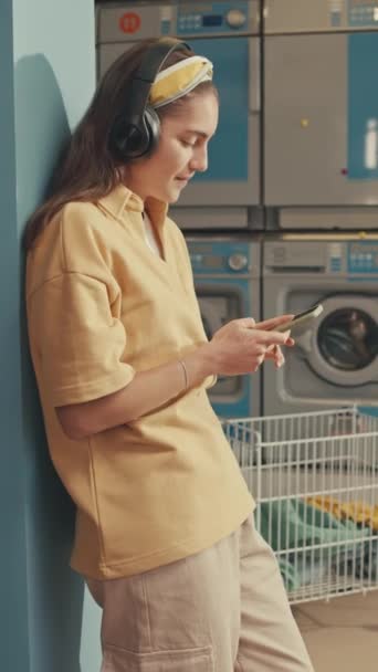 Vertical side thigh up footage of pretty Caucasian lady in headphones enjoying listening to music and chatting on phone while waiting at laundromat - Footage, Video