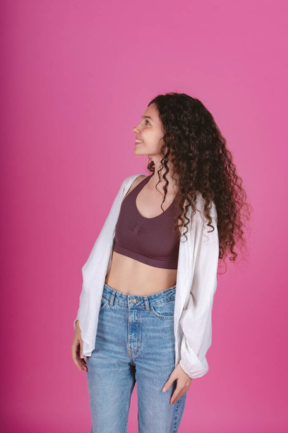 Young woman with curly hair looking upward, wearing a white cardigan and denim jeans, on a pink backdrop.  - Photo, Image
