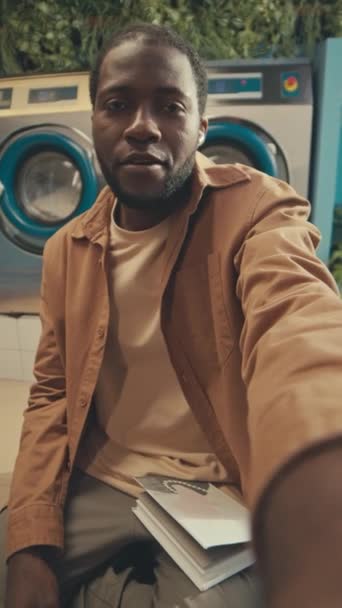 Függőleges POV UGC vidám fiatal fekete férfi forgatás vlog vagy beszél videohívás arról, hogy önkiszolgáló nyilvános mosoda - Felvétel, videó