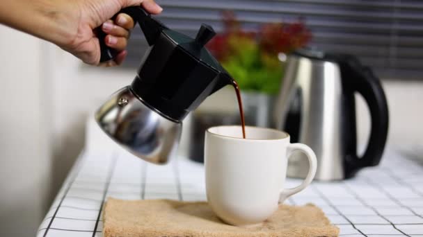 Een vrouwenhand giet zwarte koffie uit een moka pot in een witte mok, keuken interieur. Vooraanzicht, selectieve focus. - Video
