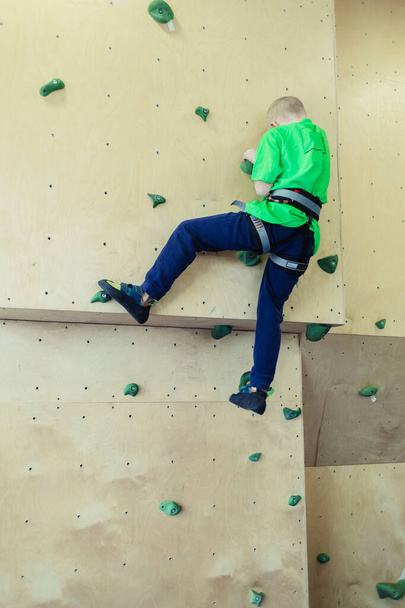 Klettern für Kinder. Blonder Junge in grünem Hemd klettert Kletterwand. Boulderkurs. Kunstrasen für sichere Aktivitäten und Wettkämpfe. Bergsteigen, Sicherheitssystem, spezielle Schuhe - Foto, Bild