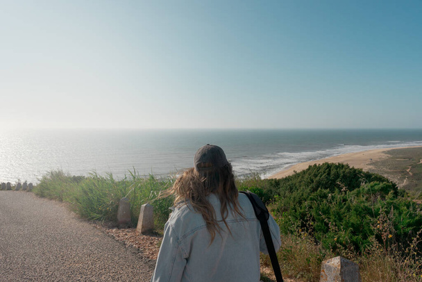 Mujer joven irreconocible caminando hacia el faro de Nazare mirando a la vista hacia Praia do Norte. Soleado día de primavera al atardecer. Vacaciones visitando el museo de surf. - Foto, imagen
