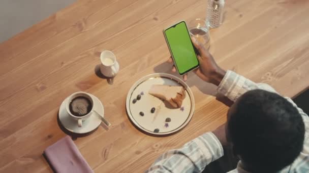 Вид зверху кадри невпізнаваного темношкірого чоловіка, який дивиться на екран ключа від зеленої хроми мобільного телефону, сидячи за столом з кавою і чізкейком в сучасному кафе - Кадри, відео