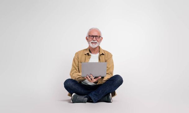 Magabiztos idősebb férfi szakmai ellenőrzése e-maileket laptop, miközben ül a fehér háttér - Fotó, kép