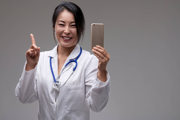 Dottoressa sorridente che tiene un telefono punti verso l'alto, suggerendo una nuova idea o funzionalità nella tecnologia sanitaria - Foto, immagini