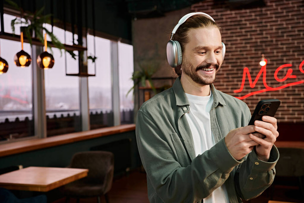 Un homme, portant un casque, regarde son téléphone dans un café moderne, plongé dans son propre monde de musique. - Photo, image