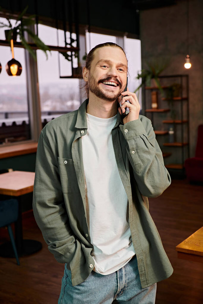 Ένας άντρας, μέσα σε ένα μοντέρνο εστιατόριο, μιλάει ζωηρά στο κινητό του ενώ στέκεται μέσα στην πολύβουη ατμόσφαιρα. - Φωτογραφία, εικόνα