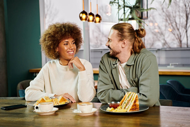 Μια Αφρο-Αμερικανίδα και ένας άντρας κάθονται σε ένα τραπέζι φορτωμένοι με πιάτα φαγητού σε ένα μοντέρνο καφέ, απολαμβάνοντας ένα γεύμα μαζί.. - Φωτογραφία, εικόνα