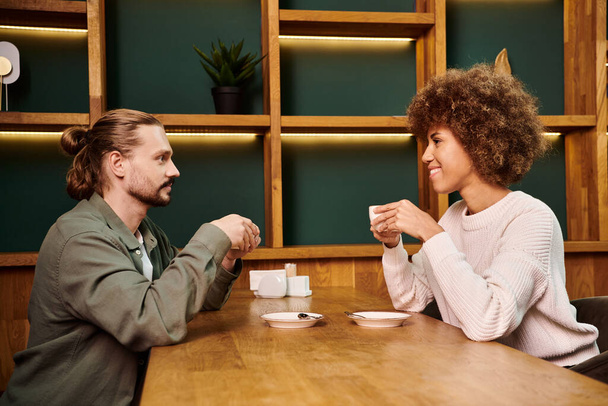 Μια Αφροαμερικανή γυναίκα και ένας άντρας κάθονται σε ένα τραπέζι σε ένα μοντέρνο καφέ, απολαμβάνοντας φλιτζάνια καφέ μαζί. - Φωτογραφία, εικόνα