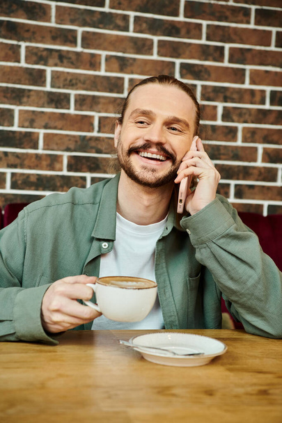 Un homme est assis à une table et engage une conversation téléphonique dans un café moderne.. - Photo, image