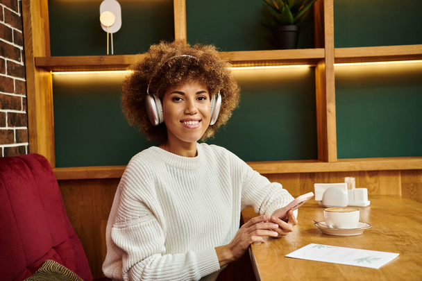 Afroameričanka sedící u stolu v moderní kavárně, zabraná do rytmu svých sluchátek. - Fotografie, Obrázek