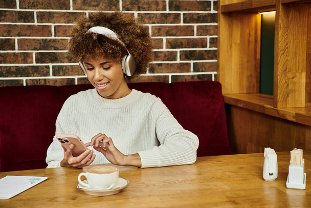 Una mujer afroamericana se sienta en una mesa en un café moderno, profundamente absorta en su teléfono celular. - Foto, Imagen