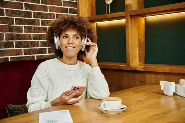 Egy afro-amerikai nő ül egy kávézóasztalnál, mobilon beszél, beszélgetésbe elegyedik. - Fotó, kép