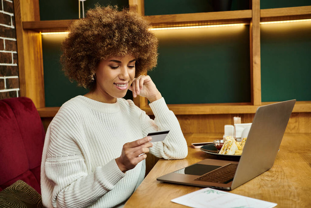 Una moderna donna afroamericana si siede in un caffè, concentrandosi intensamente sulla sua carta di credito vicino al computer portatile - Foto, immagini