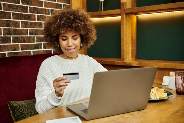 Μια σύγχρονη Αφροαμερικανίδα κάθεται σε ένα τραπέζι με ένα φορητό υπολογιστή και μια πιστωτική κάρτα, κάνοντας online αγορές. - Φωτογραφία, εικόνα